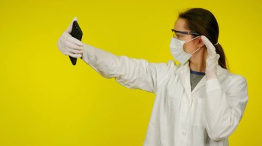 Koruyucu tıbbi maskeli, gözlüklü, eldivenli doktor akıllı telefondan selfie çekiyor.