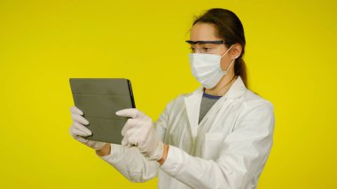 Tıbbi maskeli, gözlüklü ve lateks eldivenli genç doktor tablete bakıyor.