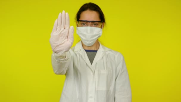 Kobieta w płaszczu medycznym, maska ochronna trzyma rękę z przodu na żółtym tle — Wideo stockowe