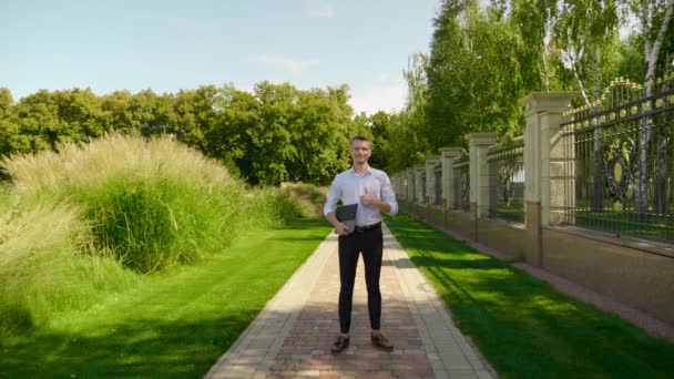 L'uomo è in piedi in un parco con un computer portatile in mano, sorridente e mostrando come — Video Stock