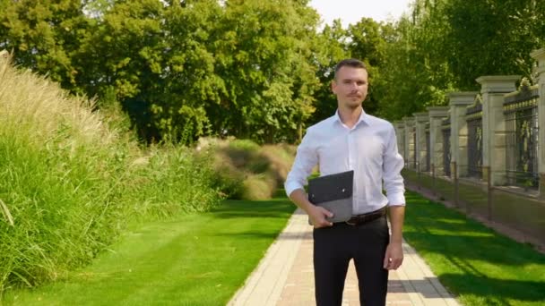 Geschäftsmann steht mit Laptop in der Hand in einem Park und blickt in die Kamera — Stockvideo