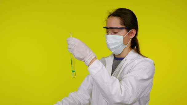 Mujer con abrigo médico, máscara protectora y gafas gotea reactivo en el tubo de ensayo — Vídeo de stock