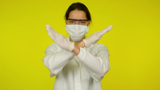Γυναίκα σε ιατρικό παλτό, προστατευτική μάσκα κρατά τα χέρια σταυρωμένα σε κίτρινο φόντο — Αρχείο Βίντεο