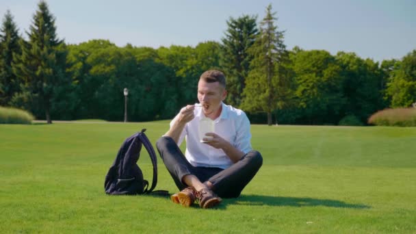 男は緑の芝生の上の公園に座って菜食主義者のサラダを食べ、それを元に戻す — ストック動画