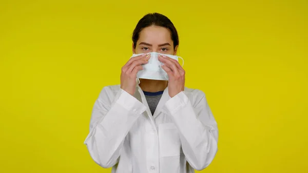 Jong meisje in een witte jas zet op een beschermende medische masker op haar gezicht — Stockfoto