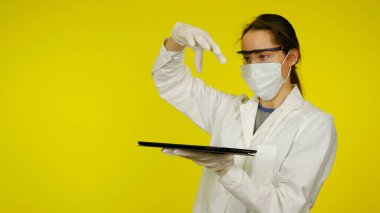 Tıbbi maskeli, gözlüklü ve lateks eldivenli doktor elinde bir tablet tutuyor.