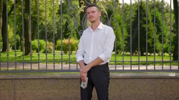 Zakenman in een wit shirt en broek staat in een park met een bundel geld — Stockvideo