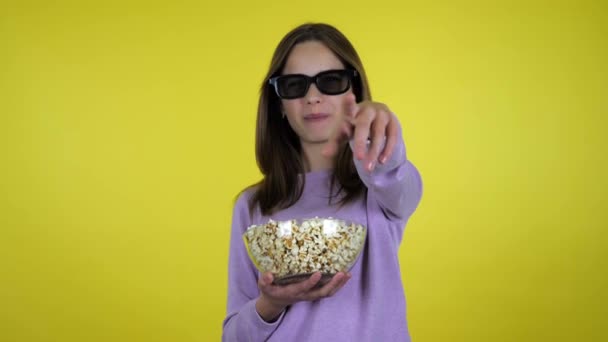 Девушка смотрит кино в черных 3D очках, ест попкорн из стеклянной чаши, смеется — стоковое видео