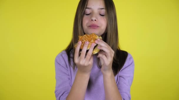 Nastolatka w różowym swetrze gryzie i zjada burgera na żółtym tle — Wideo stockowe