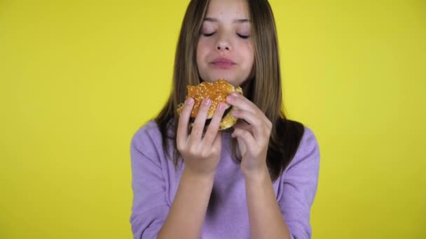 Tiener meisje in een roze trui beten en eet hamburger op gele achtergrond — Stockvideo