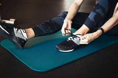 Sonra pratik egzersiz ve mavi yoga mat crossfit eğitim spor ayakkabılarını enlacing genç çekici esmer kız. Arka plan üzerinde bulanık kadındır ve dumbell odak ve ön plan. 