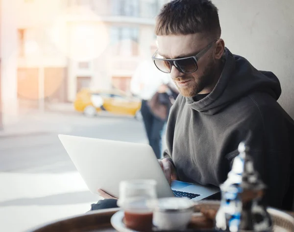 Joven pensativo sosteniendo el ordenador portátil de rodillas y trabajando en ello en la cafetería . — Foto de Stock