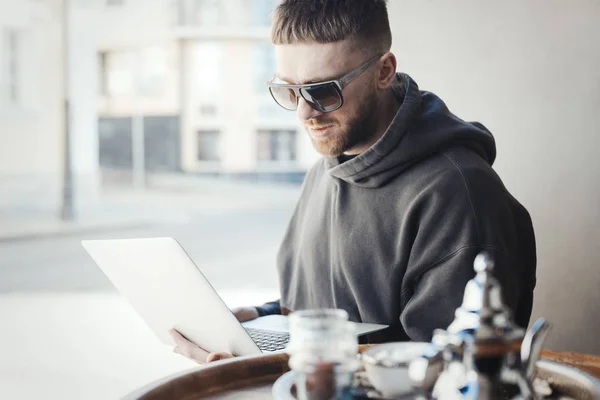 Young man in sun glasses holding laptop on his knees and working on it in cafe.