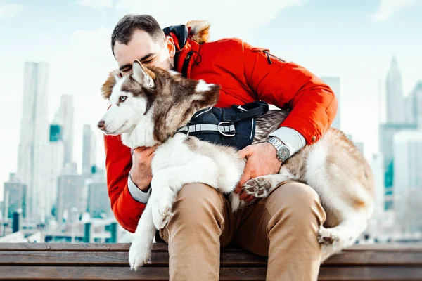 Man and Cute Siberian Husky dog sitting on bench on cityscape background. Human and animal pure Love. Best friend. — Stock Photo, Image