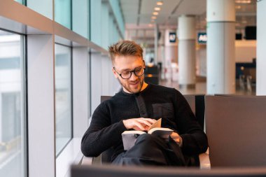 Havalimanında yolcu beklerken gözlüklü gülümseyen yolcu kitap okuyor. Başarılı iş seyahati.