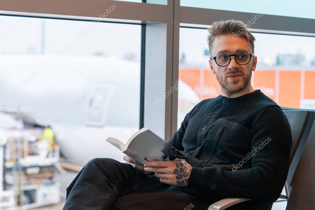 Handsome man with glasses looking at boarding information desk and holding a book while waiting for air jet at airport.
