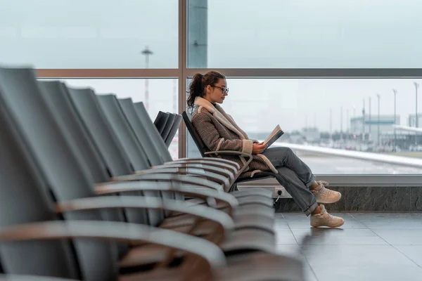 Joven mujer hermosa con gafas leyendo un libro mientras espera el embarque en Air Jet en el aeropuerto —  Fotos de Stock