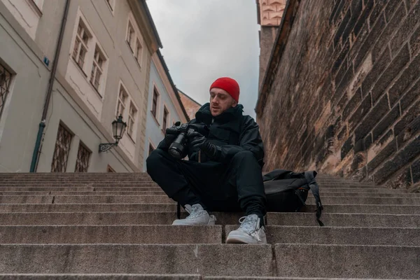Atrakcyjny mężczyzna siedzący na schodach w centrum starego europejskiego miasta i trzymający aparat fotograficzny. Współczesny stylowy Blogger i fotograf. — Zdjęcie stockowe