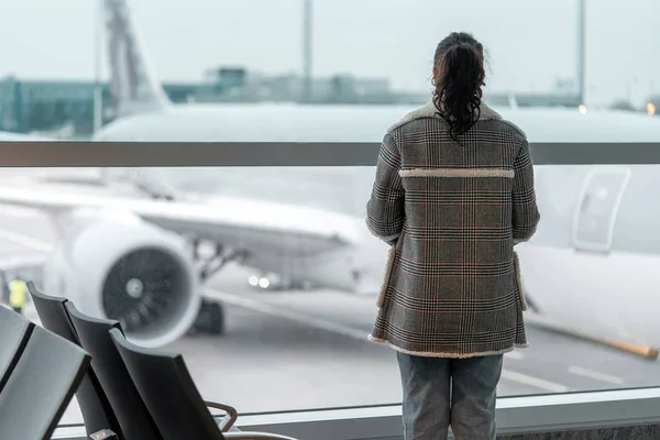 Joven morena rizado mujer cerca de gran ventana en moderno aeropuerto esperando a embarque para ir de vacaciones —  Fotos de Stock