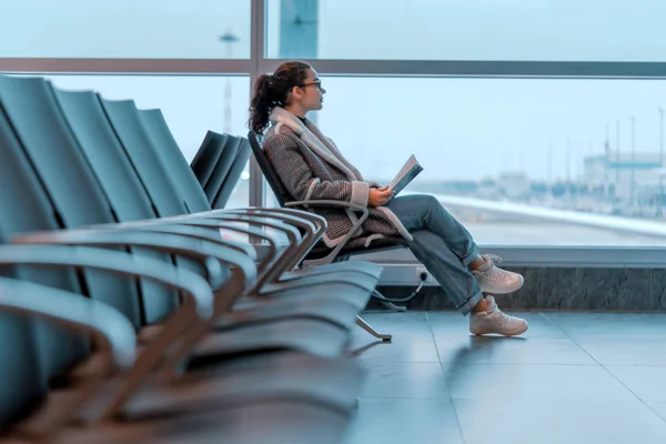 Mujer joven con gafas leyendo un libro mientras espera el embarque en el aeropuerto. El ala del avión está en el fondo y borrosa . —  Fotos de Stock