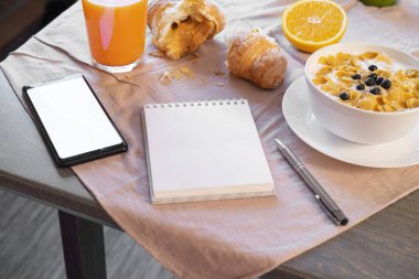 Working In Breakfast Or Brunch Time. Notepad and mobile phone with Blank screen Near Bowl with corn flakes and berries. Concept of healthy breakfast.
