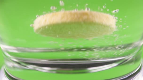 Close-up shot of An effervescent tablet against pain falls into a glass and dissolves slow-motion shot of aspirin on a green background — Stock Video