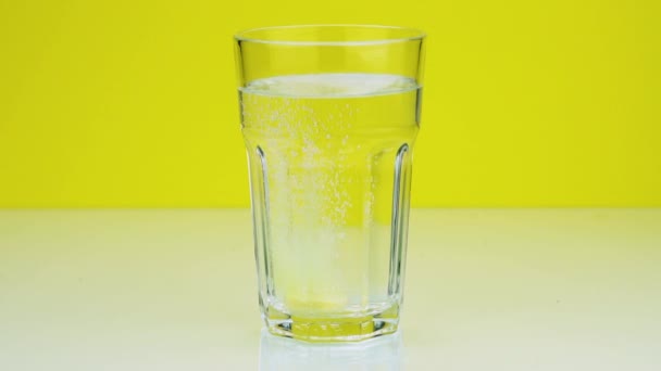Eine Brausetablette gegen Schmerzen fällt in ein Glas und löst einen Schuss Aspirin in Zeitlupe auf gelbem Hintergrund auf — Stockvideo