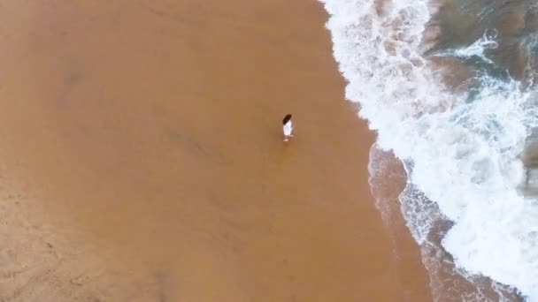 Vista aérea de una joven con un vestido blanco corto está caminando a lo largo de la playa de arena en los trópicos, cerca del océano, y moja sus pies en el agua sri lanka, hikkaduwa — Vídeo de stock