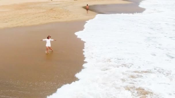 Vista superior de Uma menina em um vestido branco curto está dançando na praia de areia em hikkaduwa, sri lanka, perto do oceano, e molha os pés na água — Vídeo de Stock