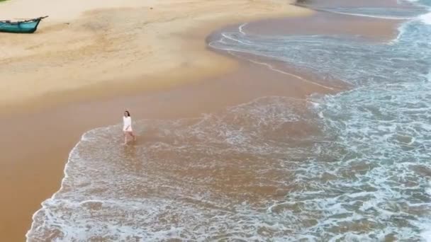 Ein glückliches Mädchen in einem kurzen weißen Kleid geht am Sandstrand in den Tropen entlang, in der Nähe des Ozeans, und benetzt ihre Füße im Wasser sri lanka, hikkaduwa — Stockvideo
