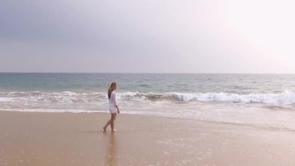 Slow motion luchtfoto A jong meisje in een korte witte jurk is wandelen langs het zandstrand in de tropen, in de buurt van de Oceaan, en haar voeten in het water wets hikkaduwa, sri lanka — Stockvideo