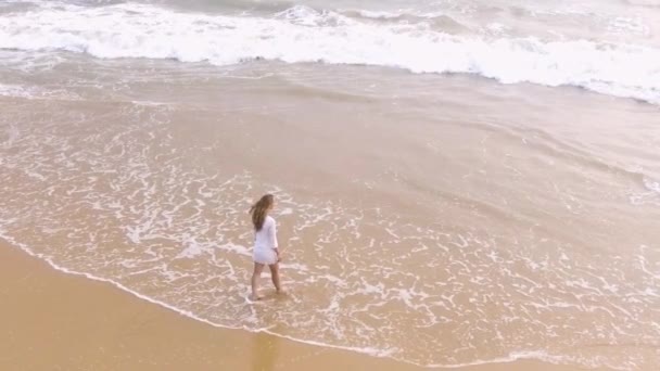 Foto aérea en cámara lenta Una joven con un vestido blanco corto está caminando por la playa de arena en los trópicos, cerca del océano — Vídeo de stock