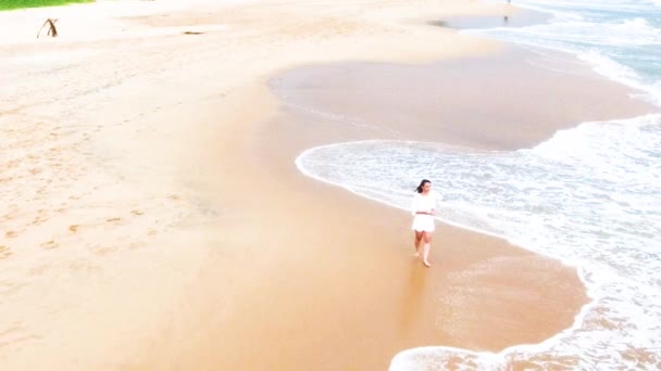 Drohnenbild eines jungen Mädchens in einem kurzen weißen Kleid läuft den Sandstrand in den Tropen entlang, in der Nähe des Ozeans, und benetzt ihre Füße im Wasser sri lanka, hikkaduwa — Stockvideo