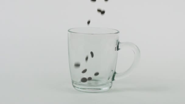 Los granos de café vuelan rápidamente, taza de vidrio transparente, fondo blanco. Concepto matutino — Vídeos de Stock