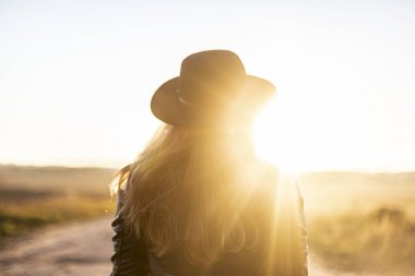 Güneşli, uzun saçlı, uzun şapkalı bir kız gün batımını seyrediyor. Arka taraf