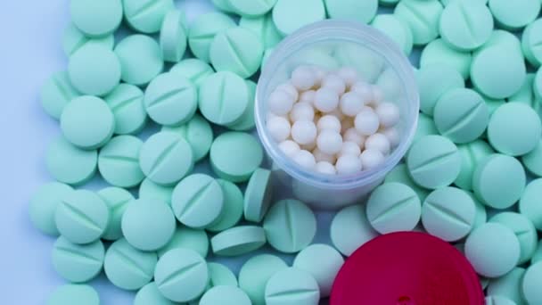 Un frasco de gránulos blancos está sobre pastillas verdes esparcidas sobre una mesa blanca — Vídeos de Stock