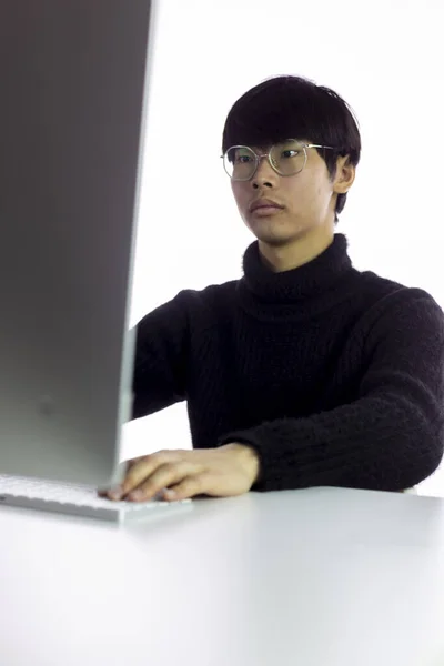 Mooie ernstige aziatische jongeman in bril werkt in kantoor op de computer — Stockfoto