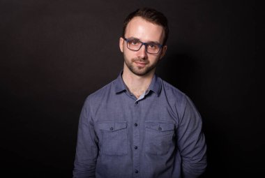 Gözlüklü çekici esmer adam kameraya özgüvenle bakıyor. Siyah arka plan