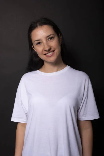 Mujer joven en camiseta blanca casual mira a la cámara con sonrisa. Fondo negro — Foto de Stock