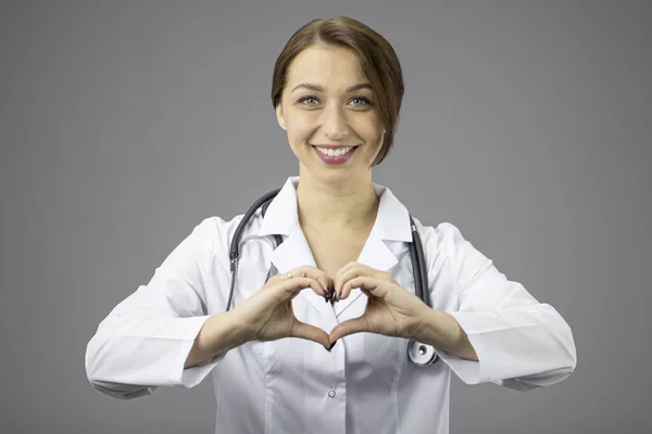 회색 배경의 섹시 한 의사는 손 과 미소로 심장을 보여 줍니다. — 스톡 사진