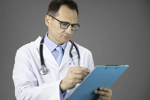 Σοβαρός γιατρός σε λευκό ιατρικό παλτό και στηθοσκόπιο γραφής σε μπλε πρόχειρο — Φωτογραφία Αρχείου