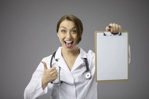 Hermoso médico ampliamente sonriendo con como signo y portapapeles en blanco en la mano —  Fotos de Stock