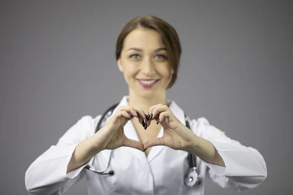 Sexy doctora en bata blanca sobre fondo gris muestra corazón y sonrisas —  Fotos de Stock