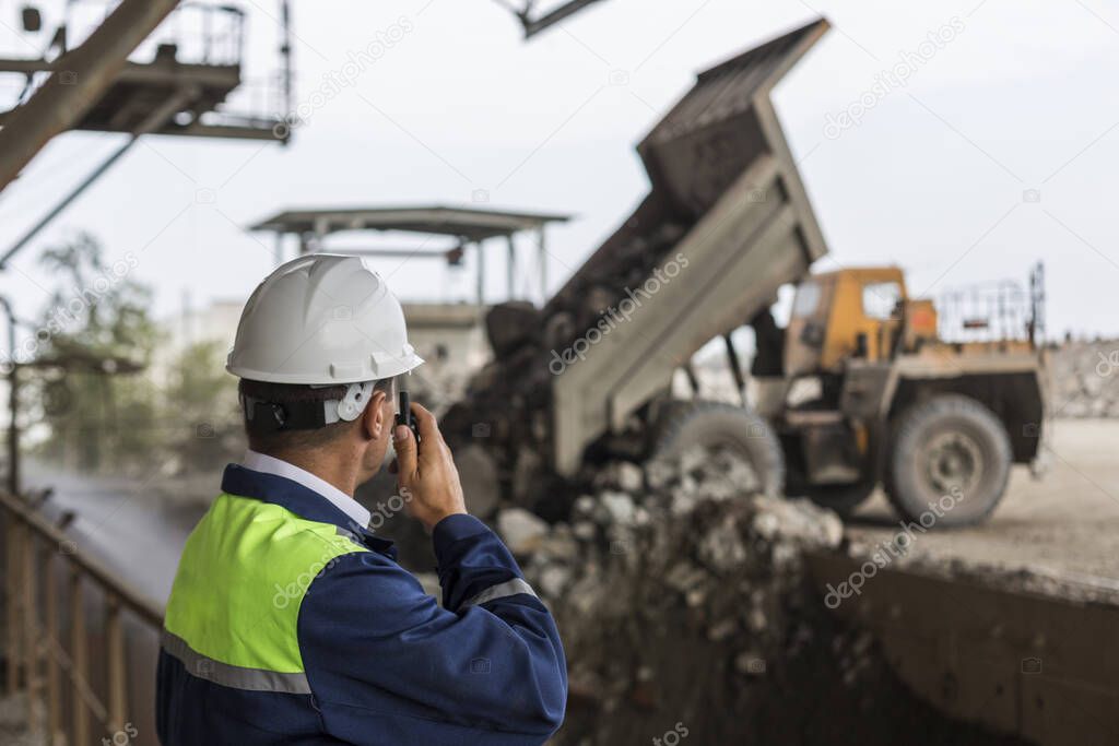 Mining engineer in yellow-blue uniform and helmet supervises unloading dumpers