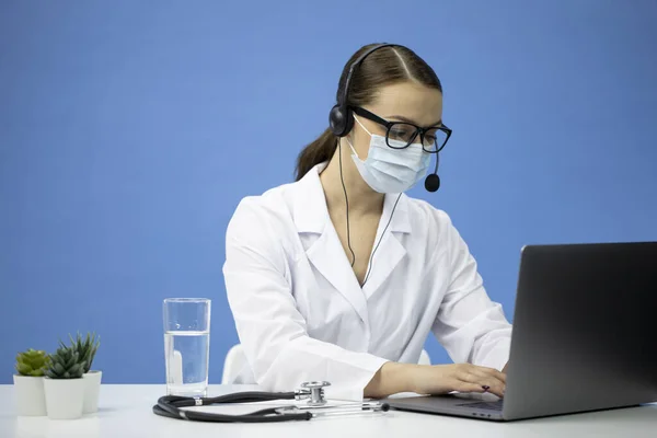 Enfermera joven en abrigo médico, máscara y auriculares da consulta en línea — Foto de Stock