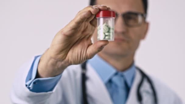 Middle aged male doctor shows transparent jar with green pills, white background — Stock Video