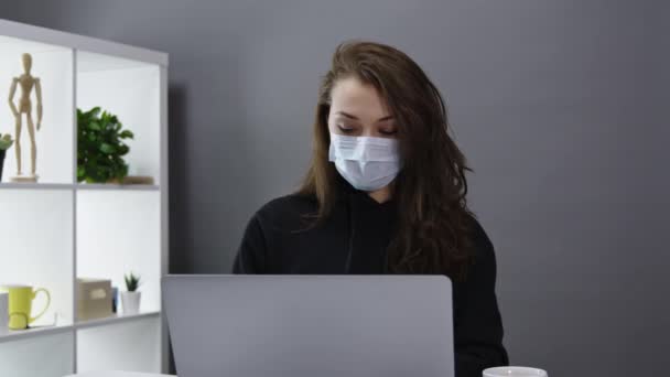 Милая деловая женщина в медицинской маске работает на ноутбуке в домашнем офисе, удаленная работа — стоковое видео