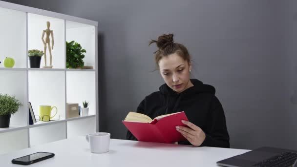 Mooi meisje in zwart hoody is geïnteresseerd lezen boek thuis en glimlachen. — Stockvideo