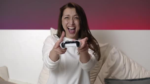 Радостная привлекательная женщина в белых пижамах играет в видеоигры с джойстиком в постели — стоковое видео
