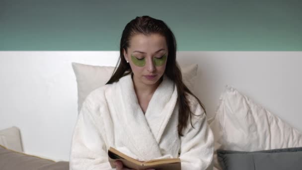 Beautiful girl in bathrobe with wet hair, patches on eyes reading book on bed — Stock Video